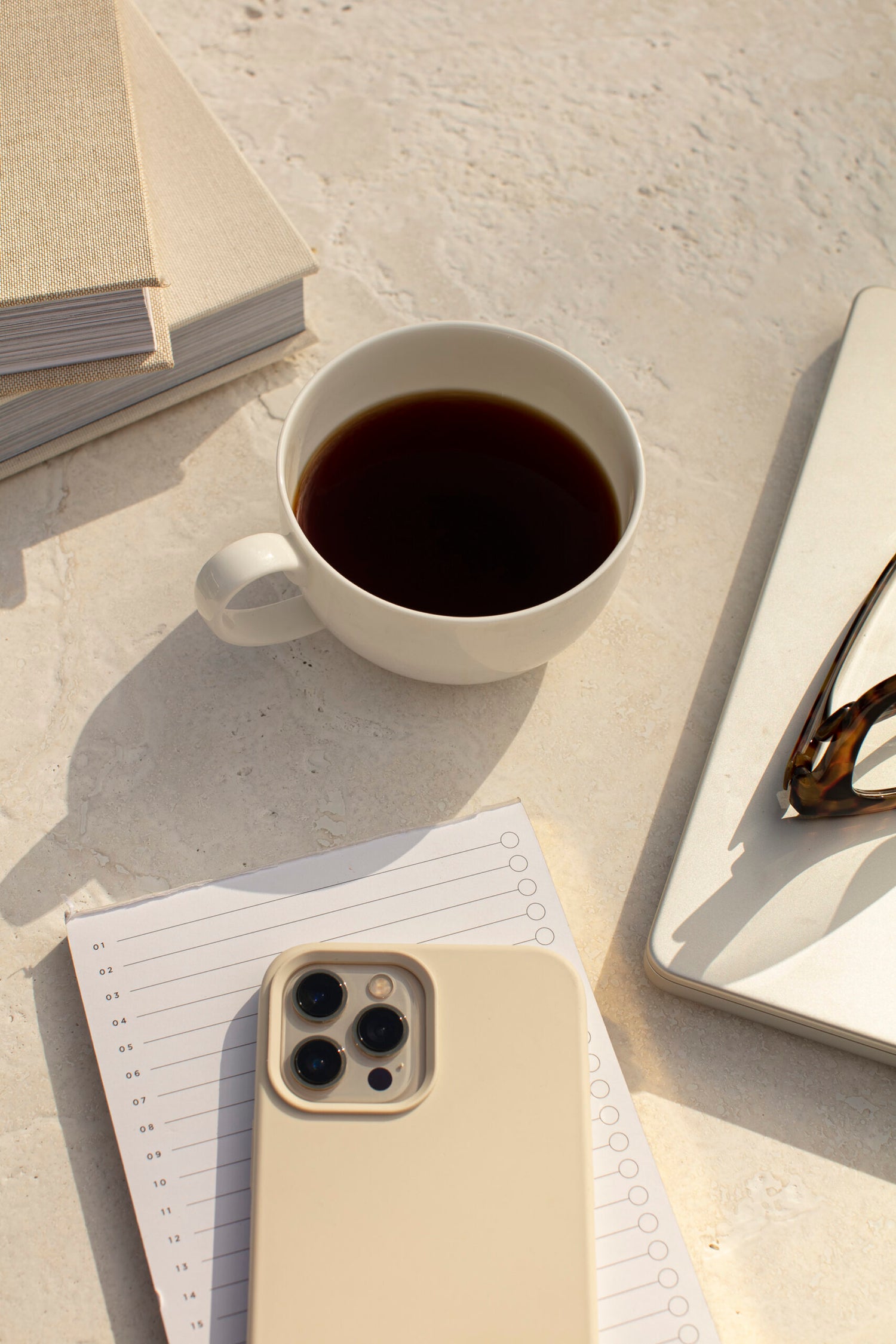 Cup of coffee on a light surface with books, phone, and sunglasses.